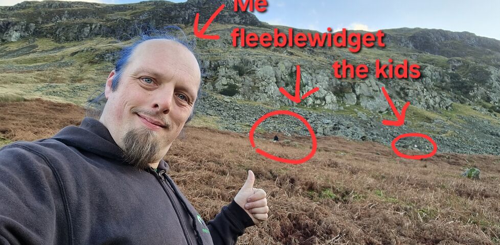 Annotated photo of Dan, a white man with blue hair, on a brackeney Lake District hillside, with arrows in the background identifying the location of 'fleeblewidget' and 'the kids'.