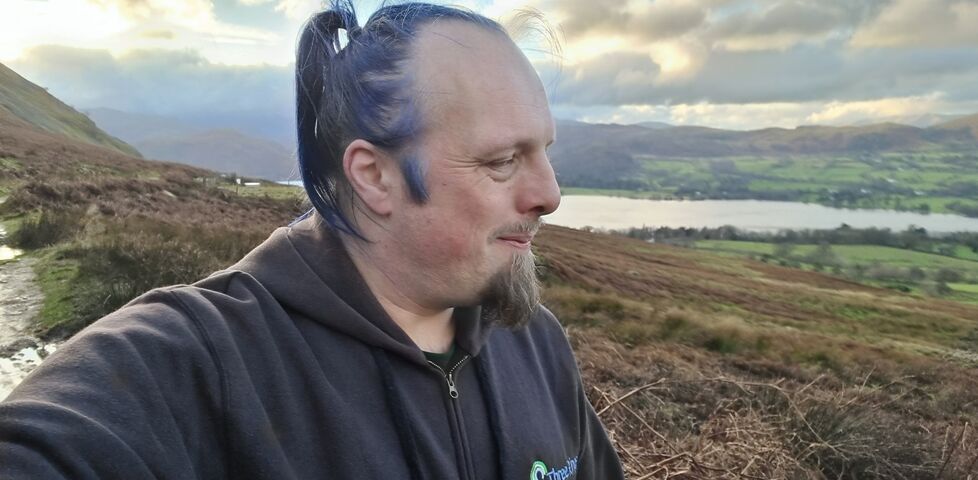 Dan, a white man with blue hair, looks over the valley of the Ullswater.