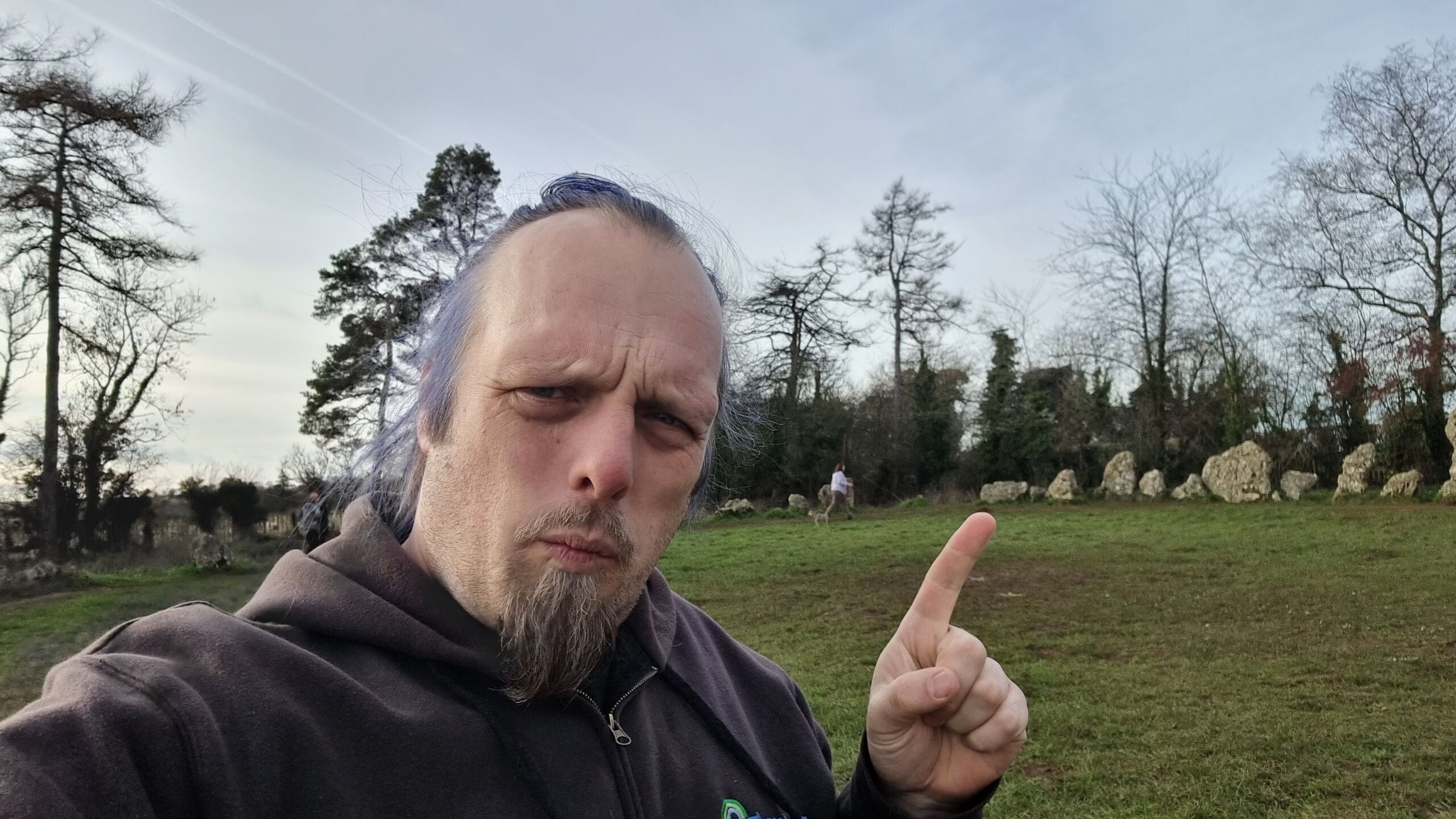 Full image of A man with blue hair points at some standing stones.