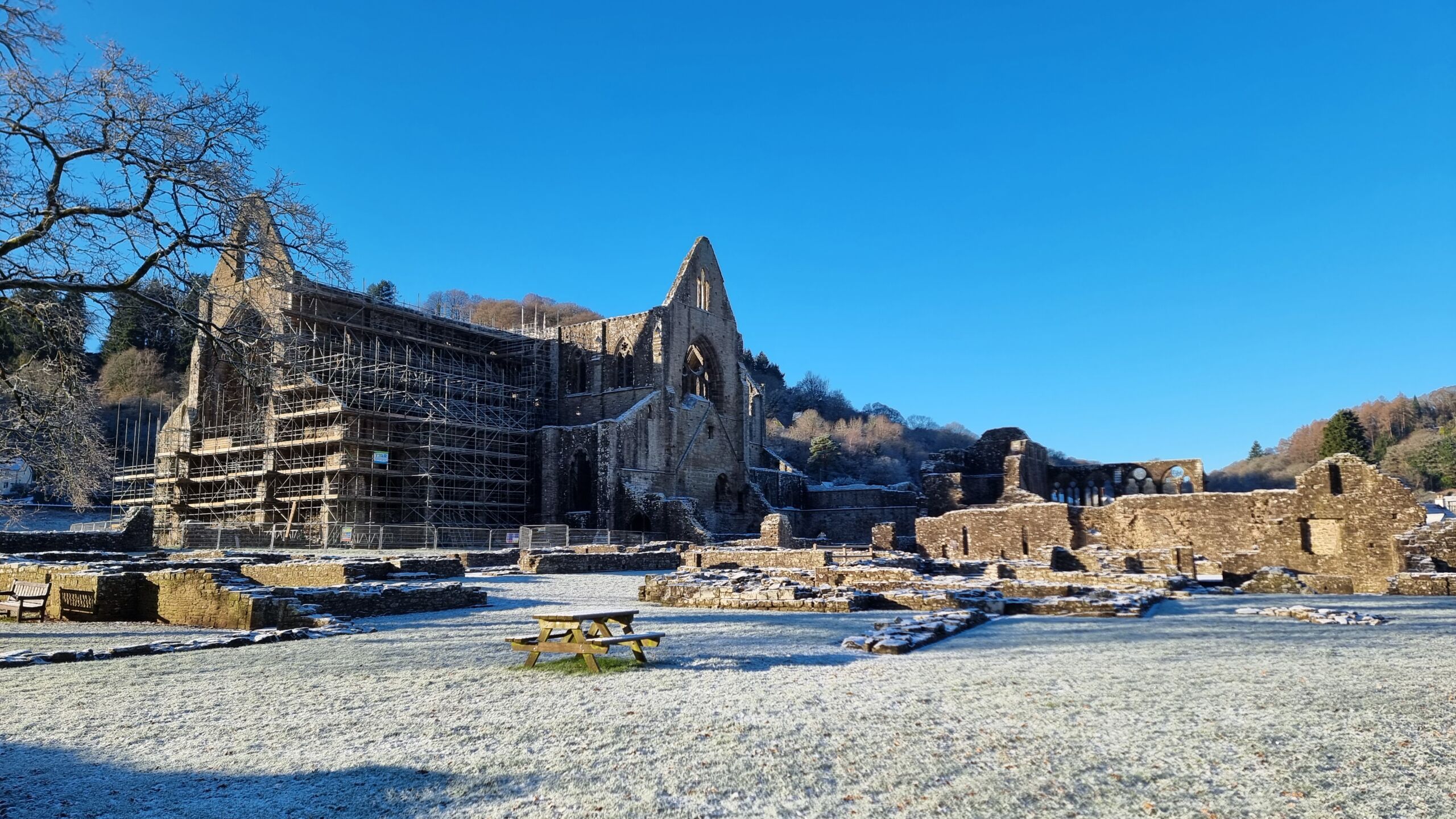 Full image of A partially-ruined stone monastery juts out of a snowy plain in a wooded valley, under bright blue skies.