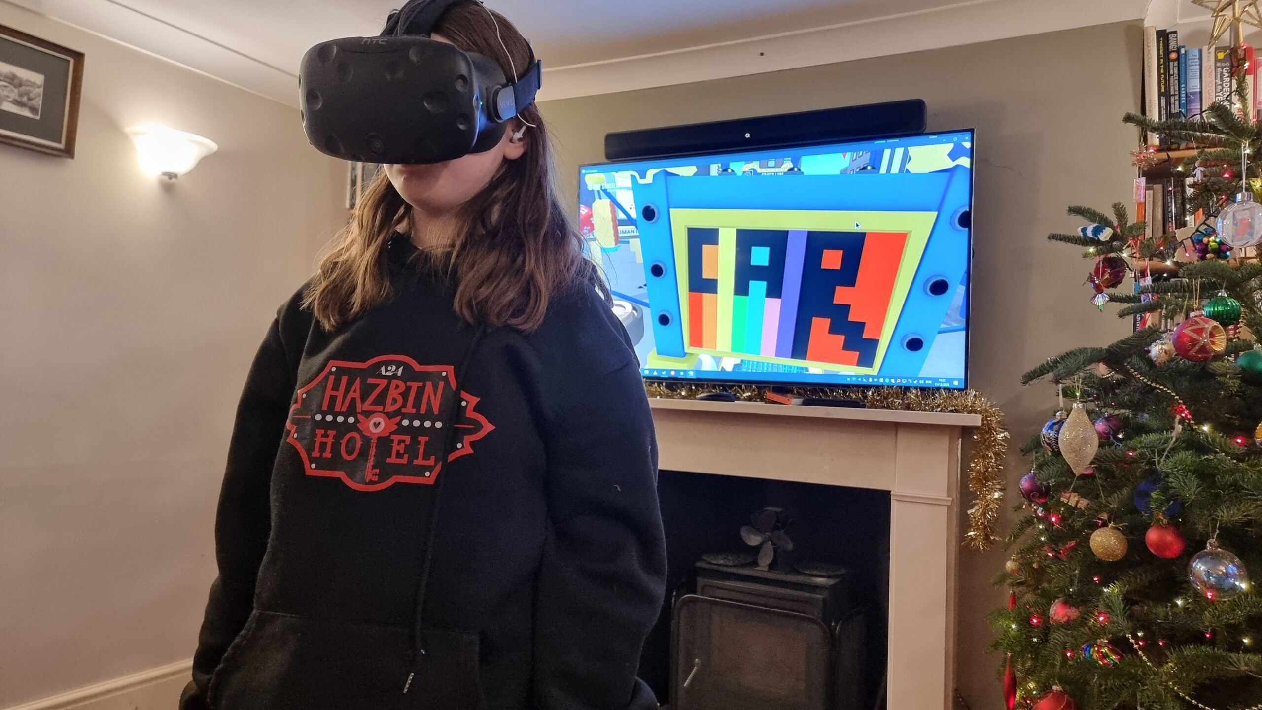 Full image of A tweenage girl in a black 'Hazbin Hotel' hoodie wears a VR headset; the screen behind her shows that she's drawn a picture featuring a rainbow background and the word 'CAR', while playing Job Simulator.