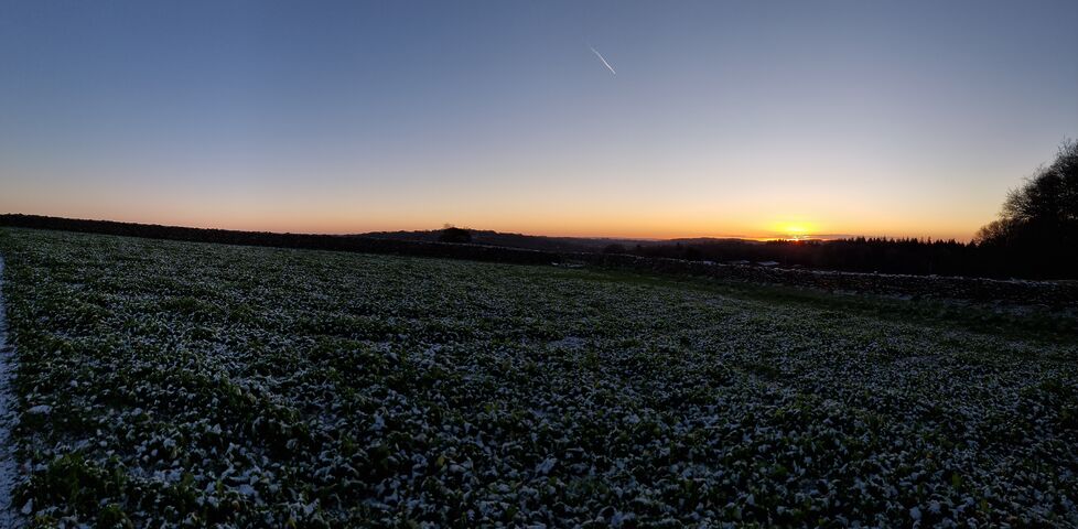 The sun peeks through clouds at the horizon beyond a wide valley full of fields.