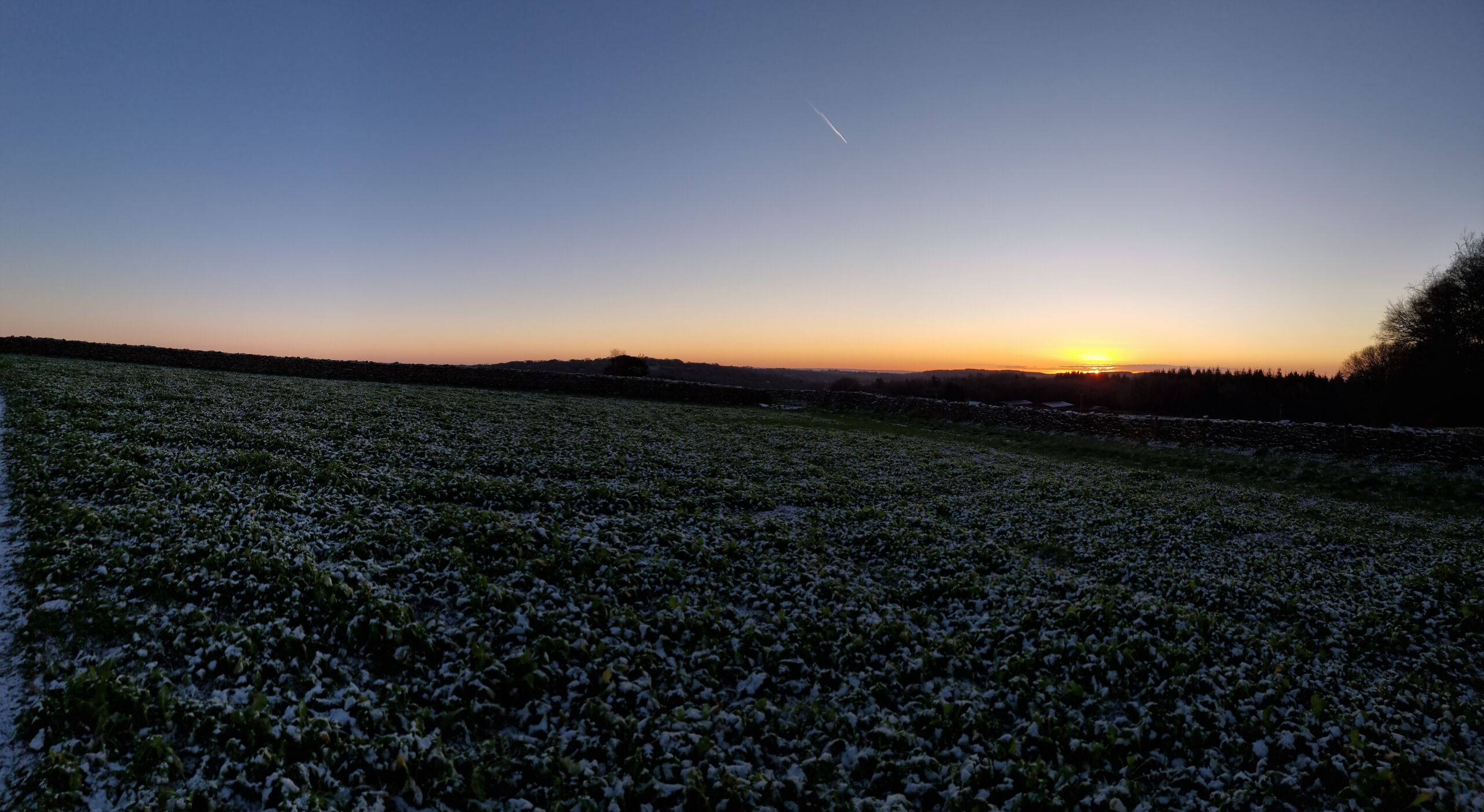 Full image of The sun peeks through clouds at the horizon beyond a wide valley full of fields.