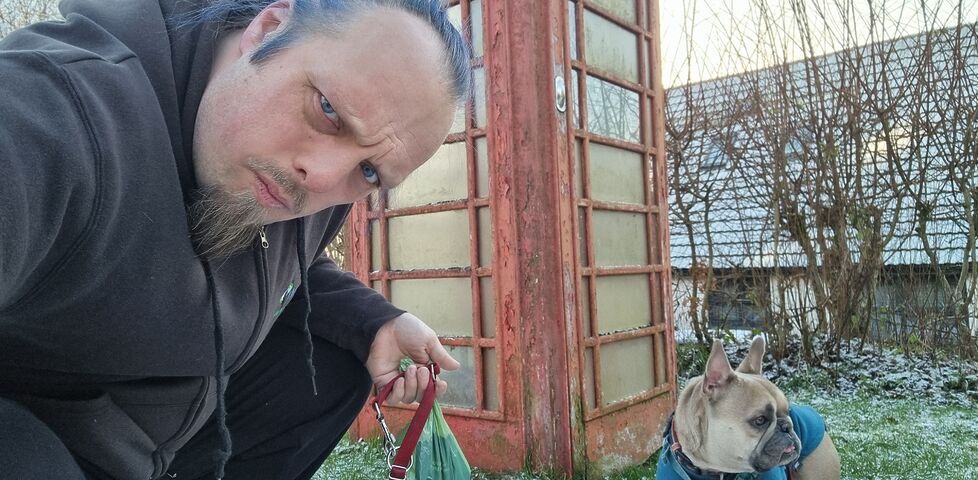 Dan, a white man with blue hair, looks confused alongside a French Bulldog, in front of a red telephone box.