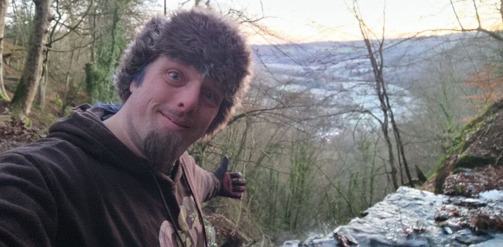 Dan, smiling and wearing a thick black hoodie and a furry hat, stands in front of a waterfall that pours into a frozen valley behind him.