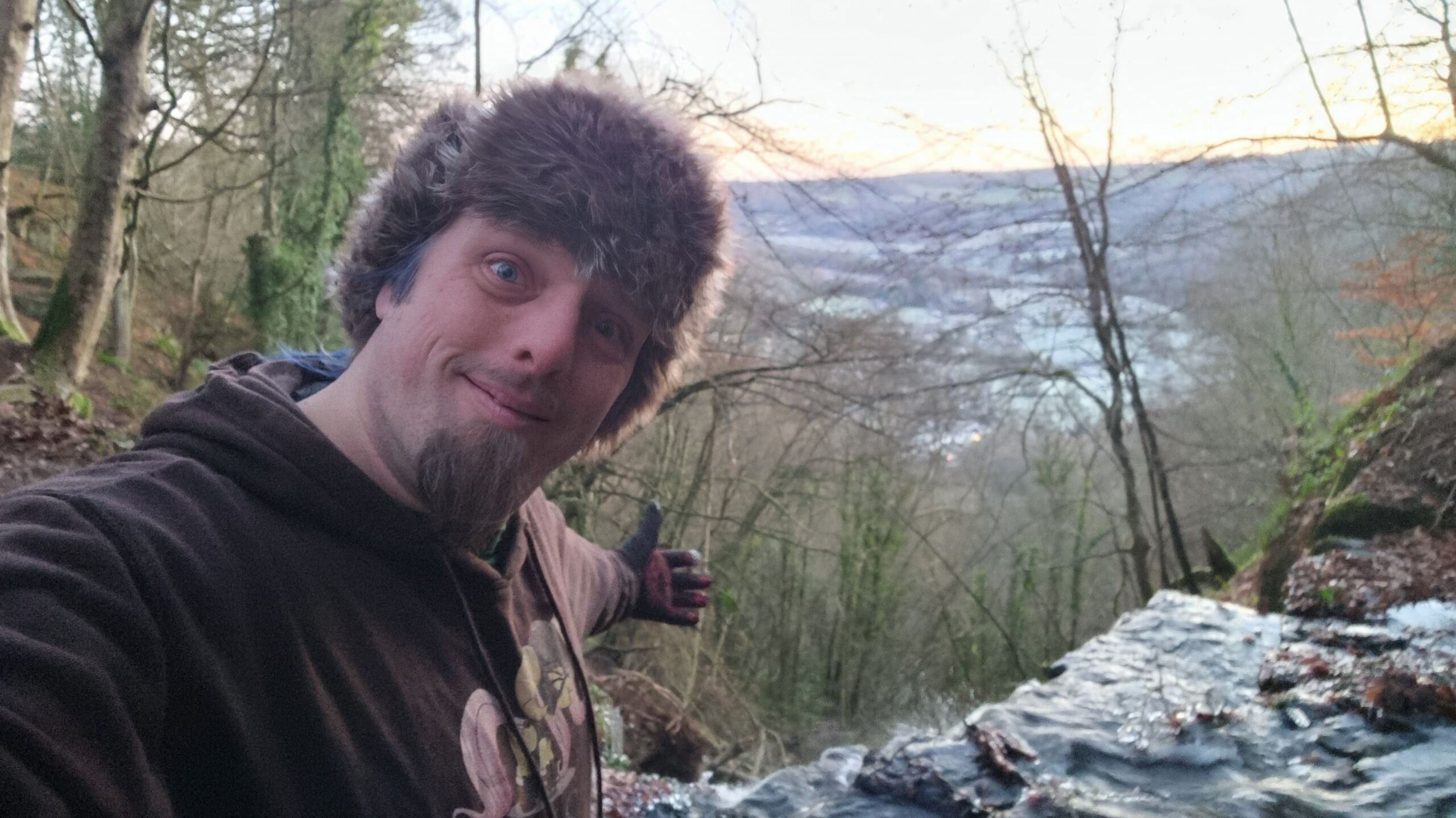 Full image of Dan, smiling and wearing a thick black hoodie and a furry hat, stands in front of a waterfall that pours into a frozen valley behind him.