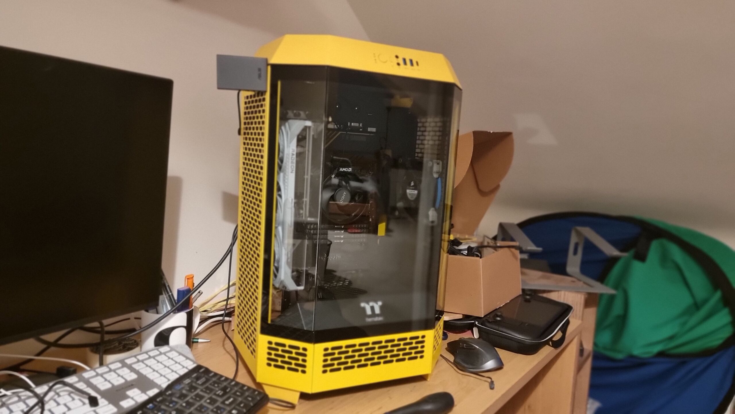 Full image of On a desk cluttered with computer parts, a partially-built PC stands in an irregular-hexagonal prism shaped case with vented yellow sides and a three-pane angled glass front.