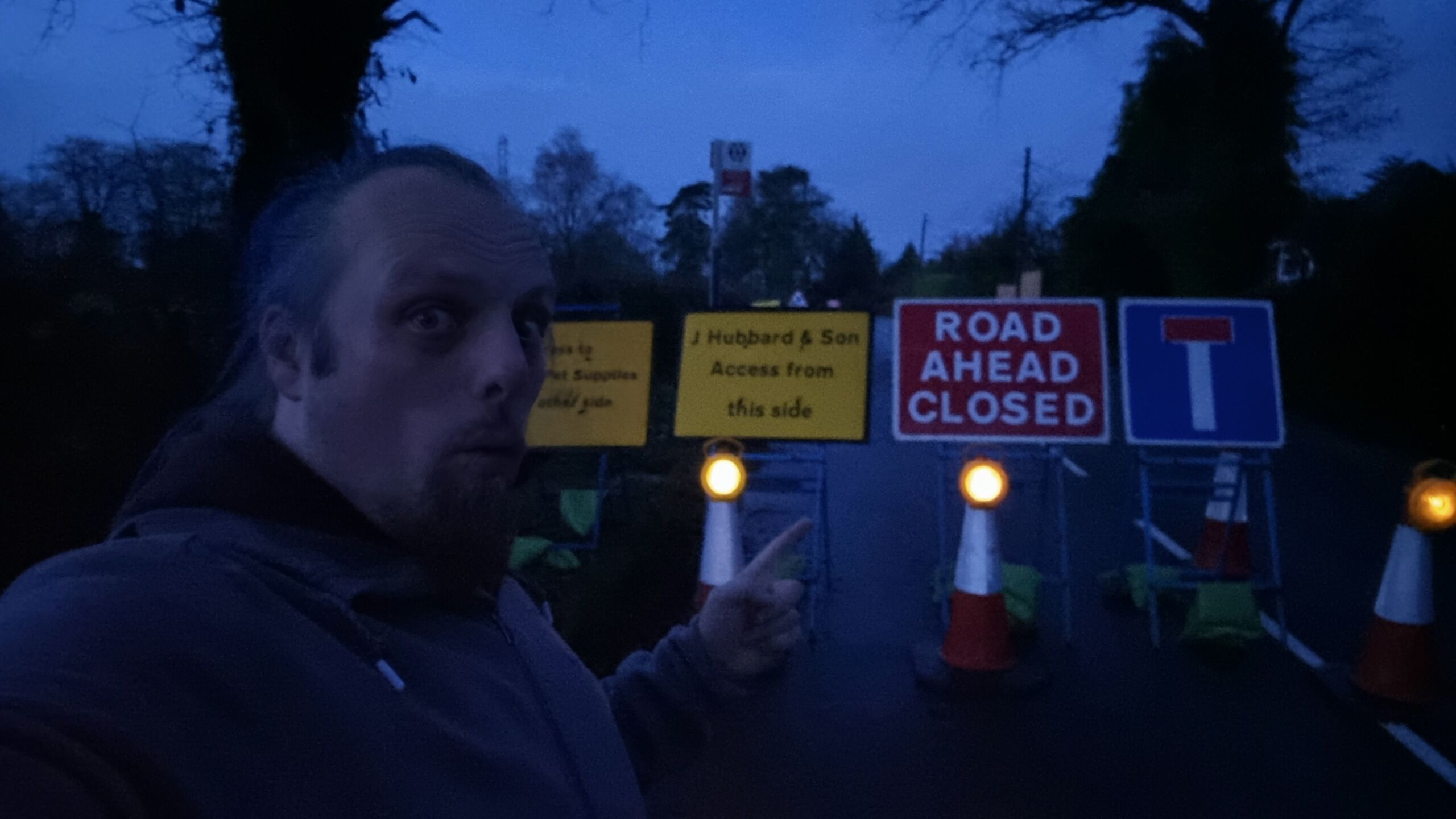Full image of Dan, in the dark, stands in front of a number of road closure signs on a rural road.