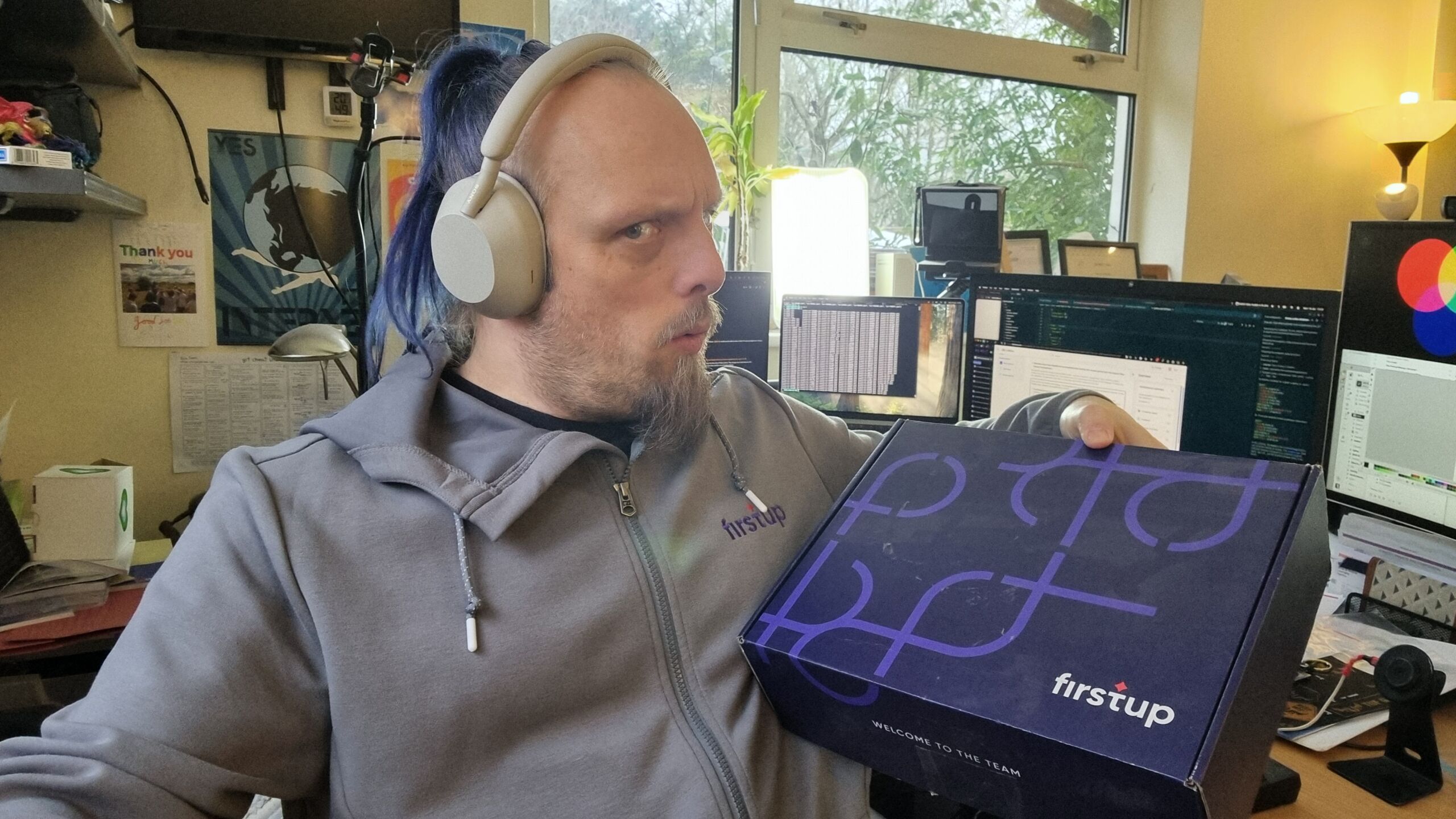 Full image of Dan, a white man with blue hair, wears headphones and a grey 'Firstup' hoodie, holding a 'Firstup'-branded shoebox.