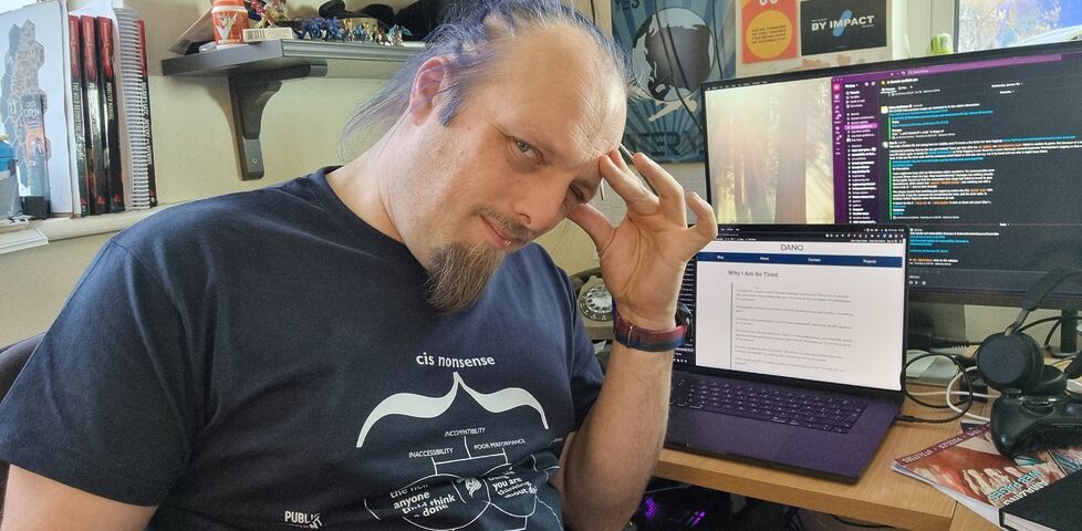 Dan, a white man with a goatee beard and long blue hair, sits at a laptop on a cluttered desk with his fingers to his temple and a pained and exhausted look on his face.