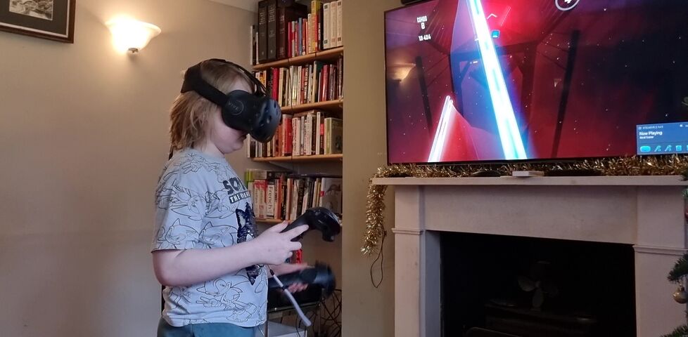 A boy wearing a white t-shirt and a VR headset holds a pair of controllers which on the screen behind him we can see appear to him as lightsabres.