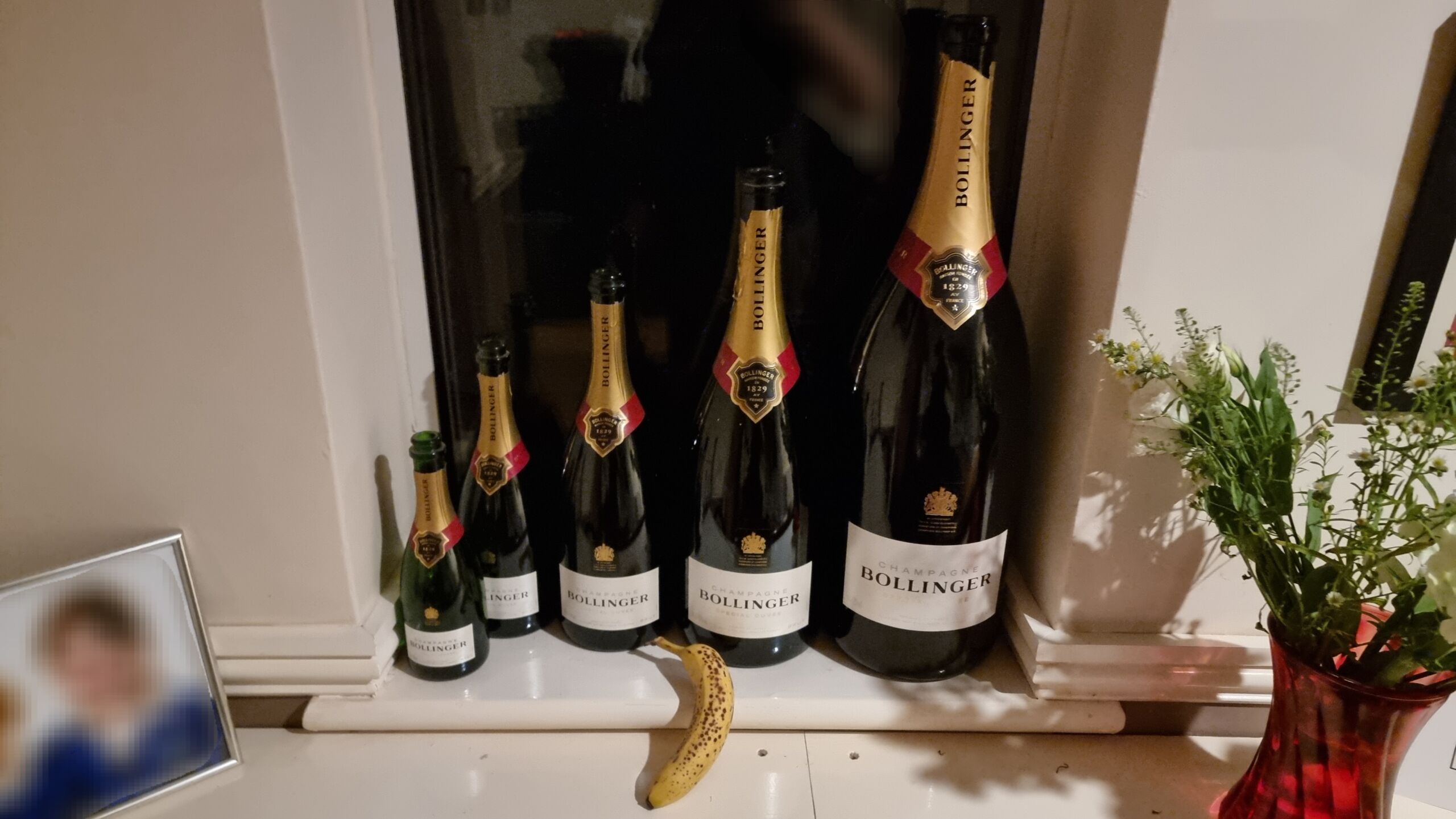 Full image of A half bottle, standard bottle, magnum, Jeroboam, and Methuselah of Bollinger champagne lined up on a window ledge, with a banana for scale.