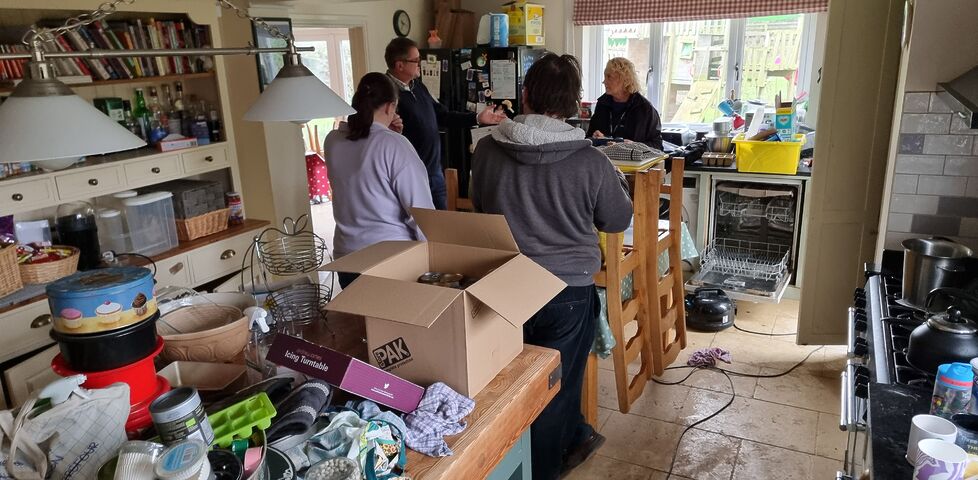 A group of adults stand talking in a disrupted kitchen, with food, furniture, and boxes stacked high.