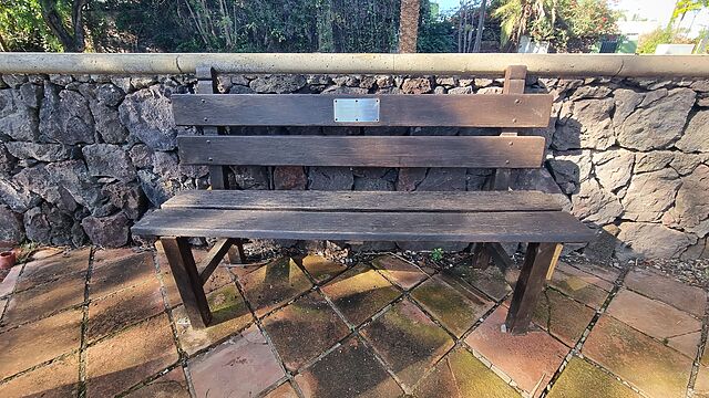On a tiled patio, a wooden bench stands, bearing a plaque that reads: THIS MEMORIAL GARDEN IS DEDICATED TO THE MEMORY OF THOSE WHO LOST THEIR LIVES IN THE AIR DISASTER IN TENERIFE ON 25TH APRIL 1980.