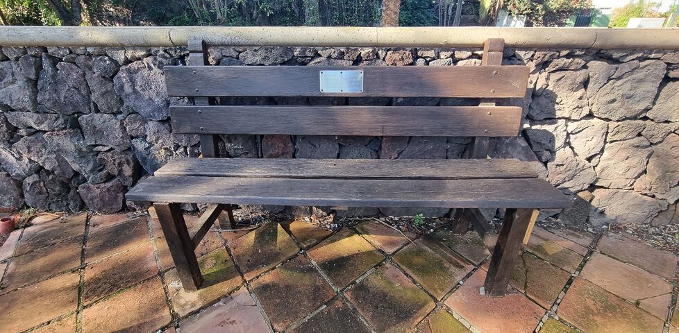 On a tiled patio, a wooden bench stands, bearing a plaque that reads: THIS MEMORIAL GARDEN IS DEDICATED TO THE MEMORY OF THOSE WHO LOST THEIR LIVES IN THE AIR DISASTER IN TENERIFE ON 25TH APRIL 1980.