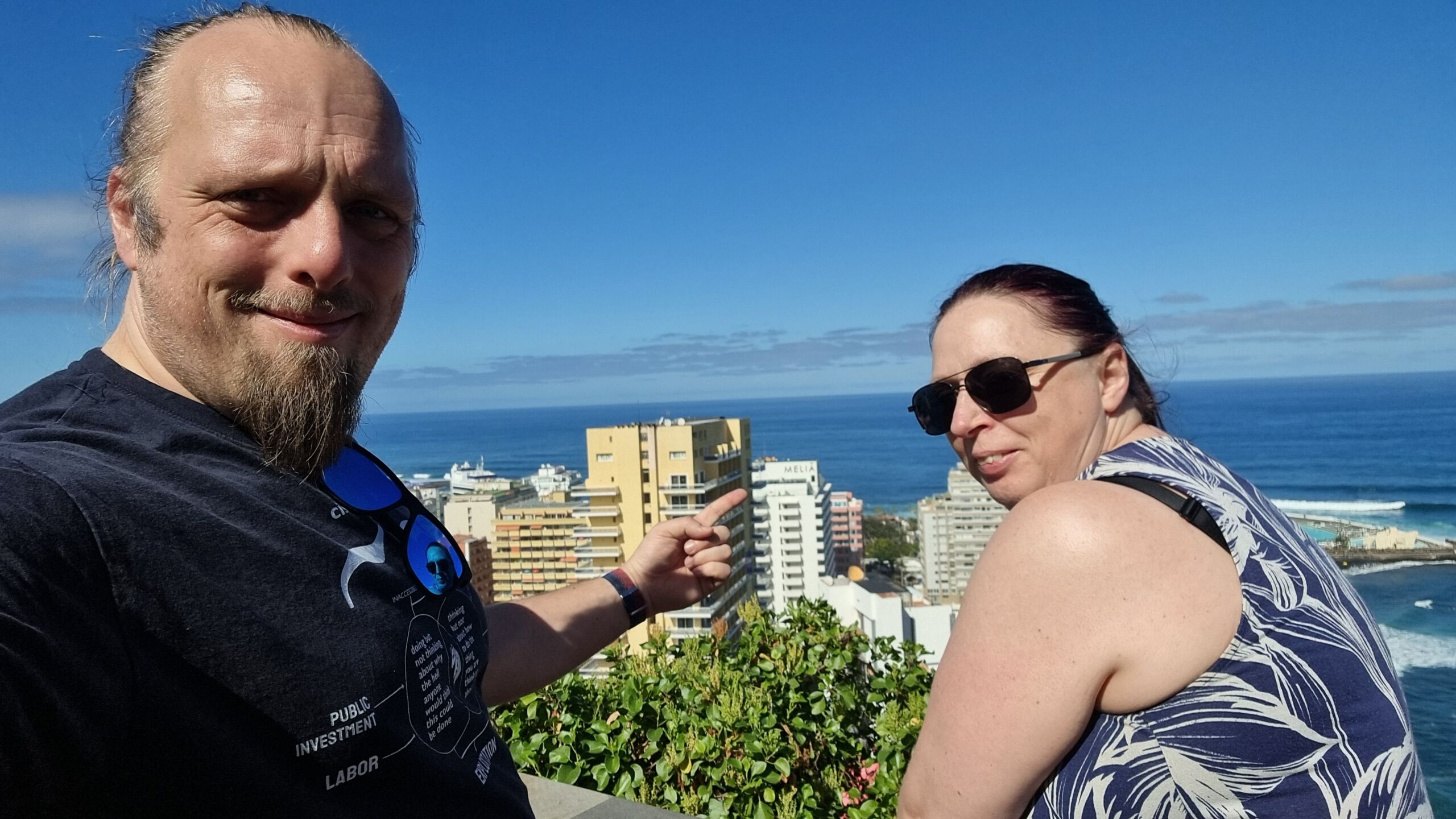 Full image of Dan and Ruth stand on an overlook, pointing at a seaside Meliá hotel in the distant city below.