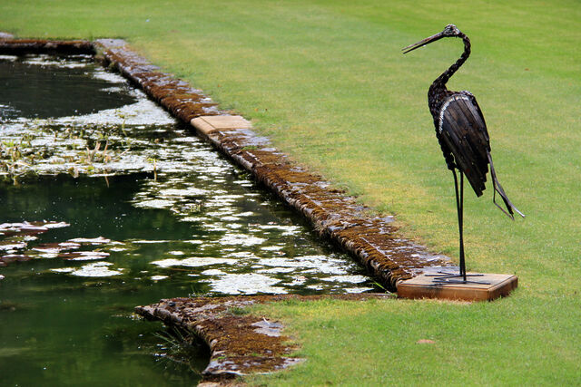 Metalwork fake heron alongside a manicured pond.