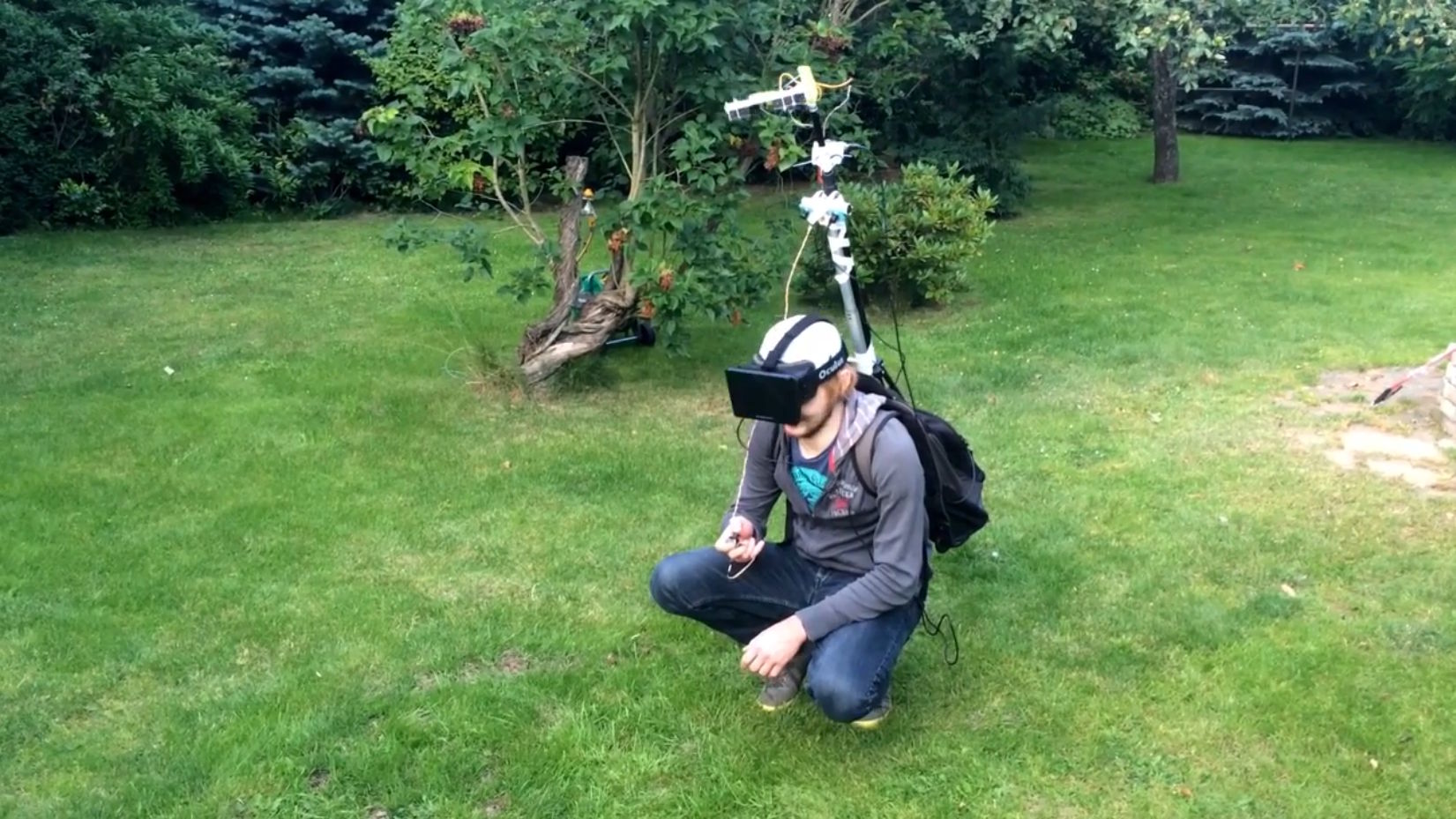 Full image of In a cultivated garden, a white man crouches, wearing an Oculus Rift VR headset connected to a backpack, from which is extended a tall pole above his head with cameras attached.