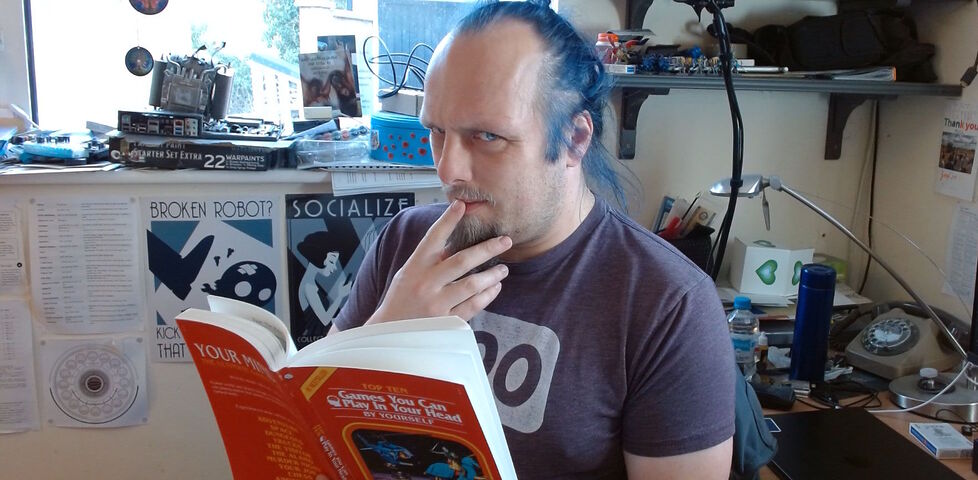 Dan, a white man with blue hair, sits in a cluttered office, a finger to his lips as he's deep in thought, reading a red book titled 'Top Ten Games You Can Play In Your Own Head By Yourself'.