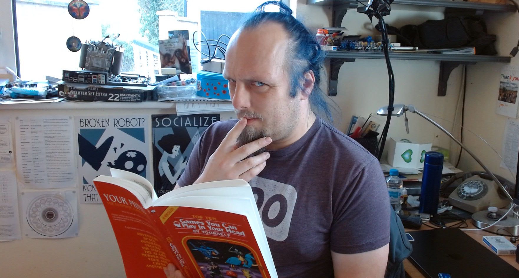 Full image of Dan, a white man with blue hair, sits in a cluttered office, a finger to his lips as he's deep in thought, reading a red book titled 'Top Ten Games You Can Play In Your Own Head By Yourself'.