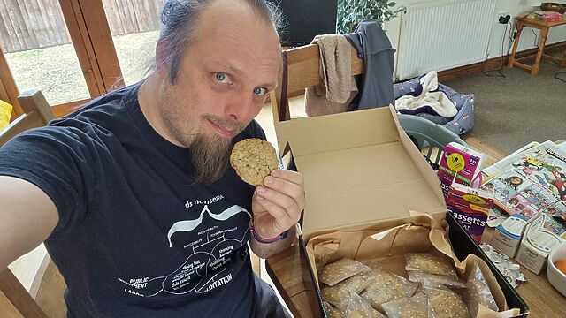 Dan, a white man with a goatee beard and blue hair, sits next to a large box of cookies, about to eat one.