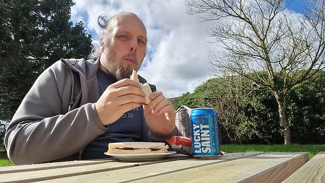 Dan, a white man with a goatee beard and long hair, sits at a wooden picnic bench in front of a sandwich, crisp packet, and can of Lucky Saint.