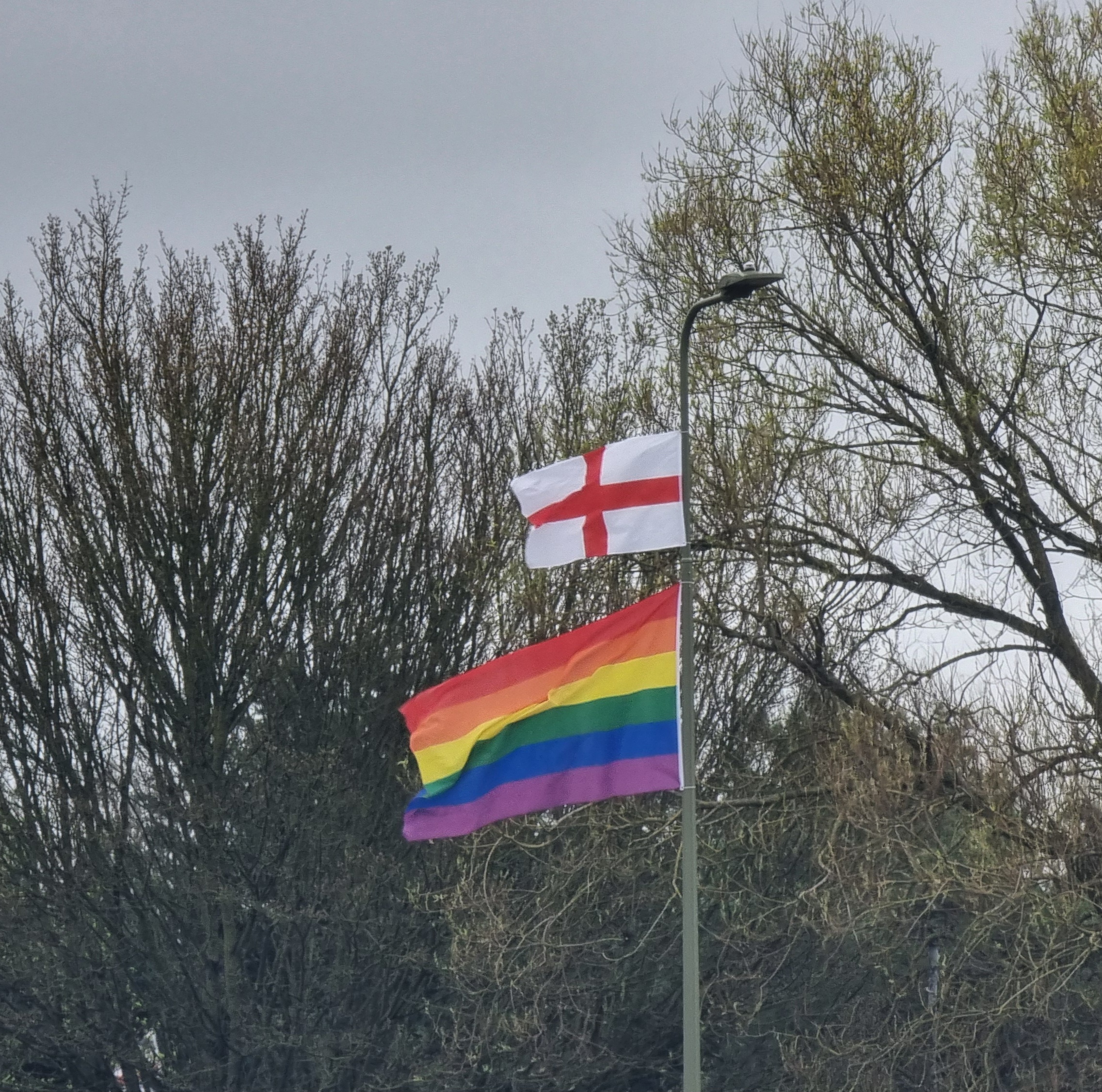 Full image of Two flags hang from a tall lamp post. The smaller upper one is a St. George's Cross, the flag of England.the lower, larger (and much nicer) one is the six-colour Pride rainbow.