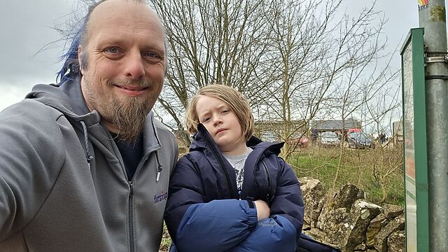 Dan and a child stand at a bus stop, with Diddly Squat Farm Shop in the background.