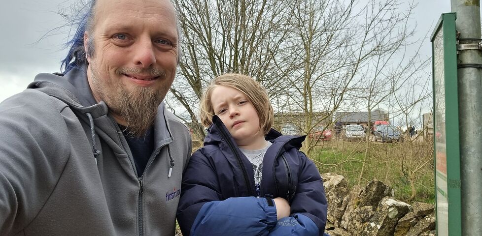 Dan and a child stand at a bus stop, with Diddly Squat Farm Shop in the background.