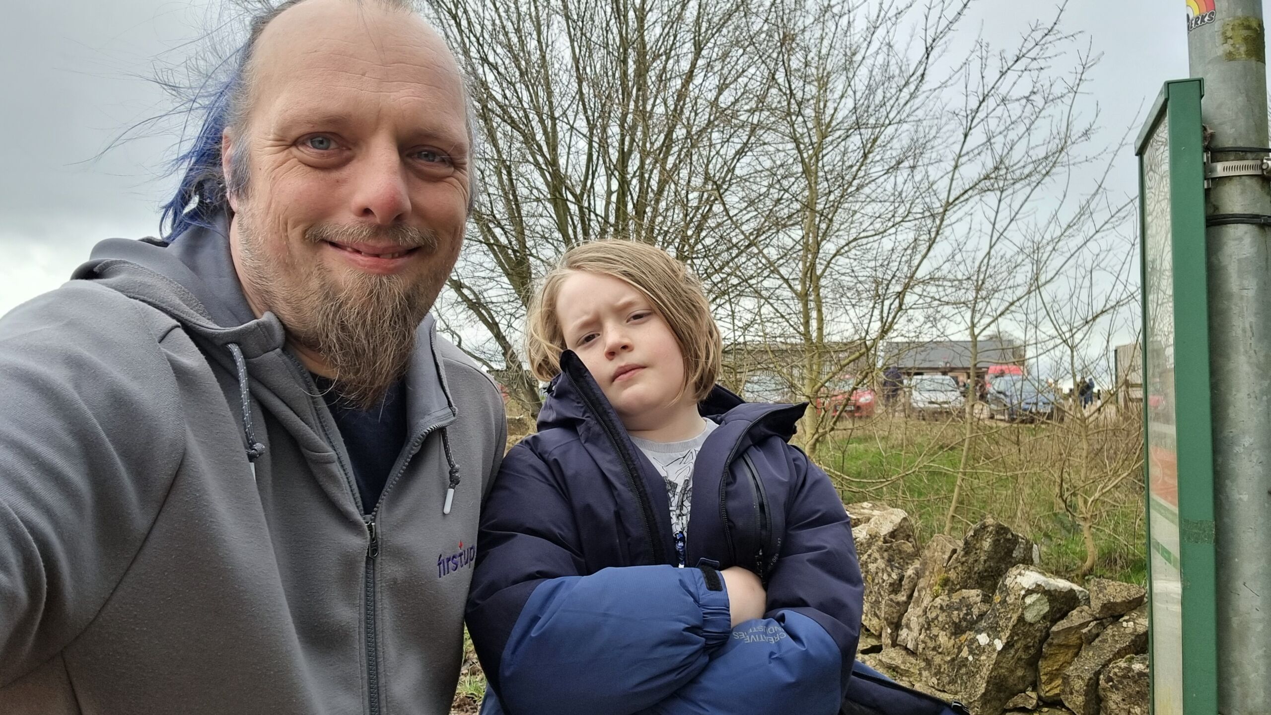 Full image of Dan and a child stand at a bus stop, with Diddly Squat Farm Shop in the background.