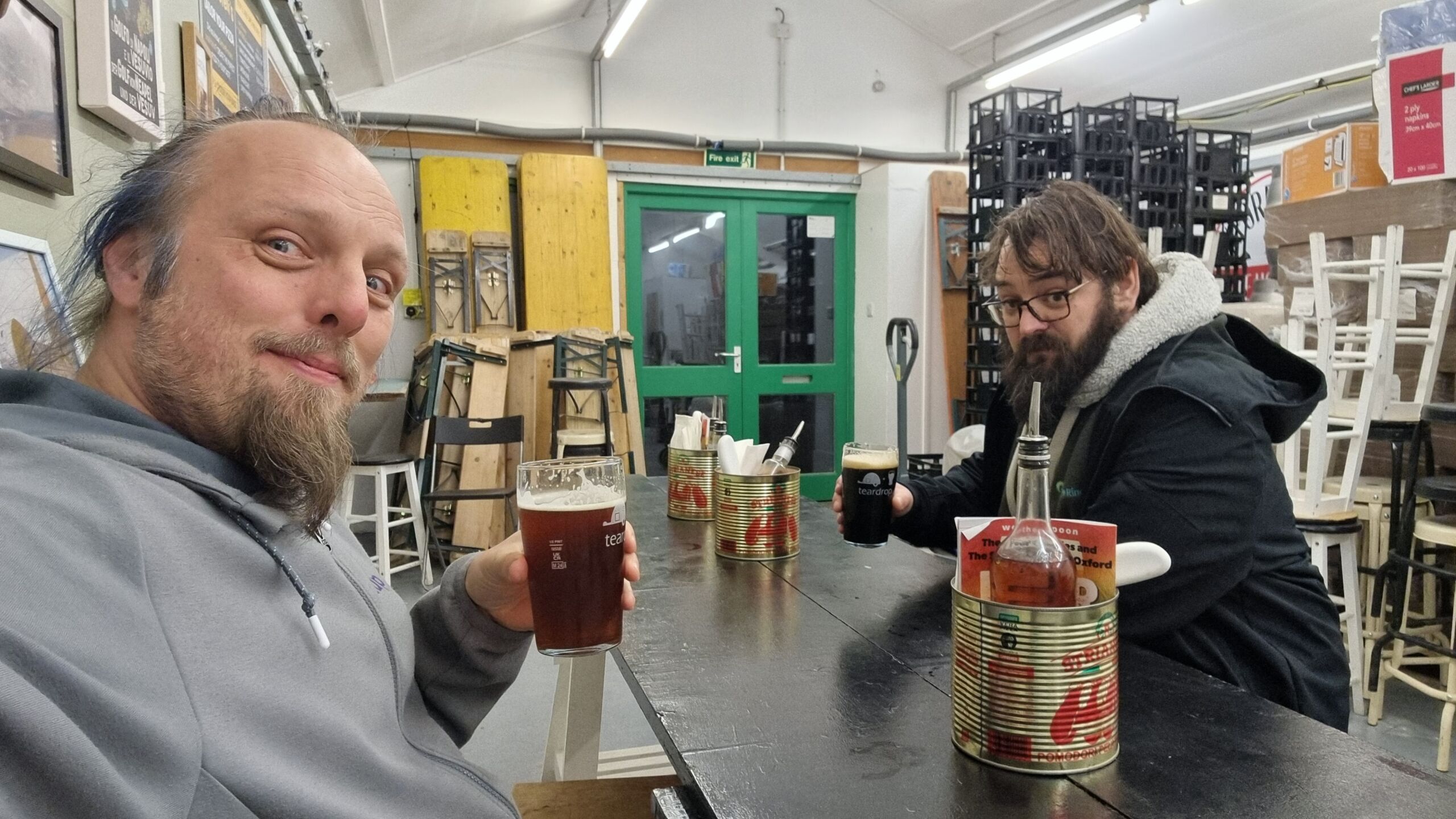 Full image of Dan and JTA sit with pints of beer in a cluttered brewery, at a table with large tomato cans repurposed as holders for chilli oil and pizza cutters.