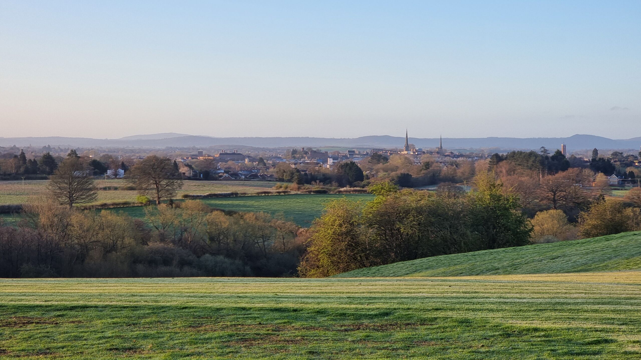 Full image of A view of Shrewsbury, from a distance, nestled in its valley, as the Spring's morning sun dapples across the verdant grass below.