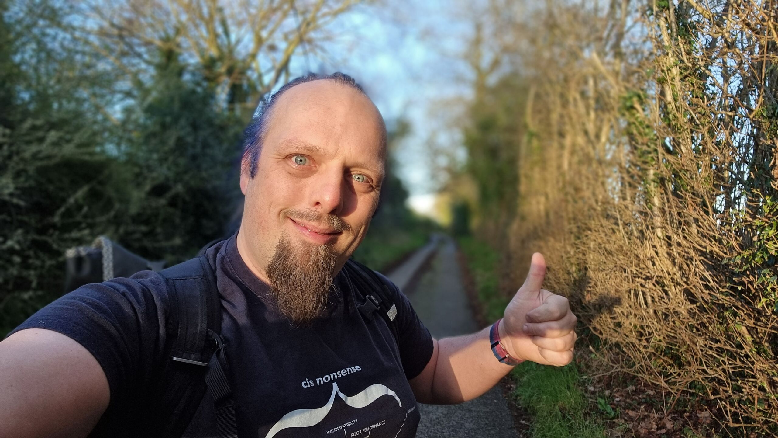 Full image of Dan throws a thumbs-up as the sun breaks through the tree-lined footpath he's walking along.