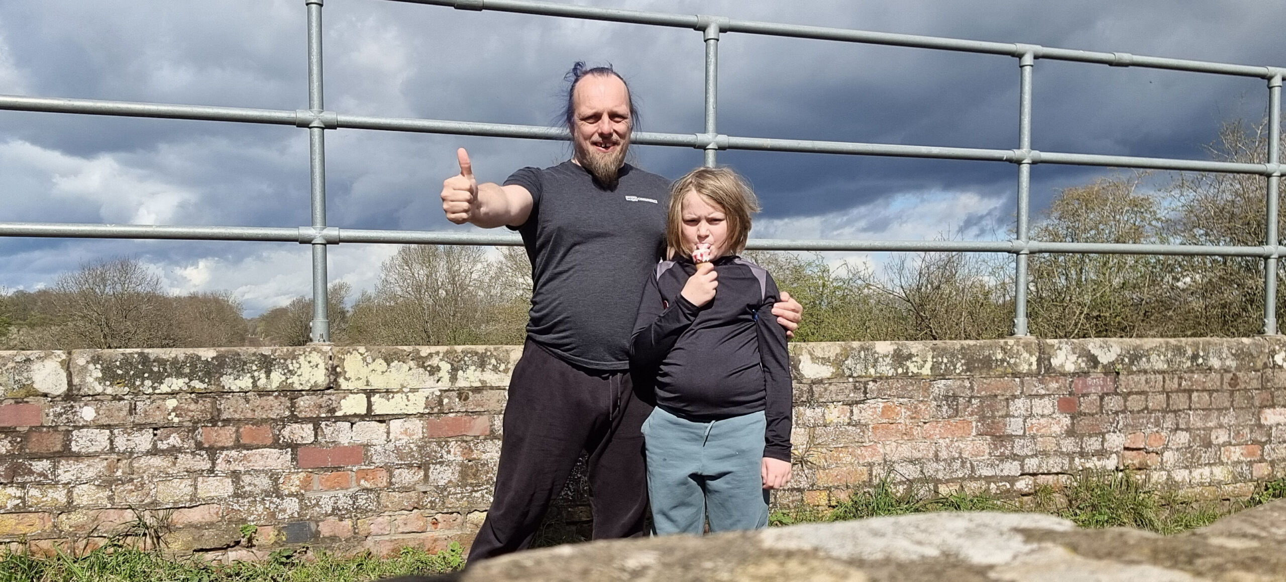 Full image of Dan and a small child throw a thumbs-up to the camera from atop a brick bridge.
