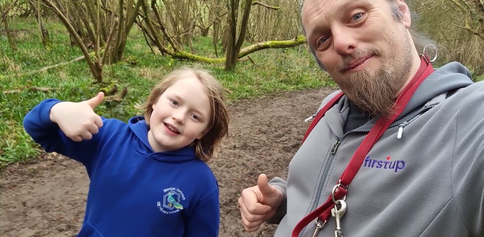 Dan and a boy each throw a thumbs-up to the camera from a forest footpath.