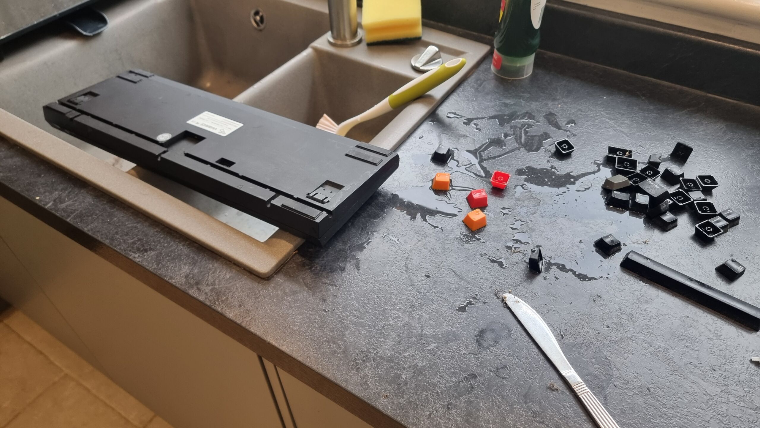 Full image of A desktop computer keyboard inverted over a kitchen sink, with many of its keycaps scattered around.