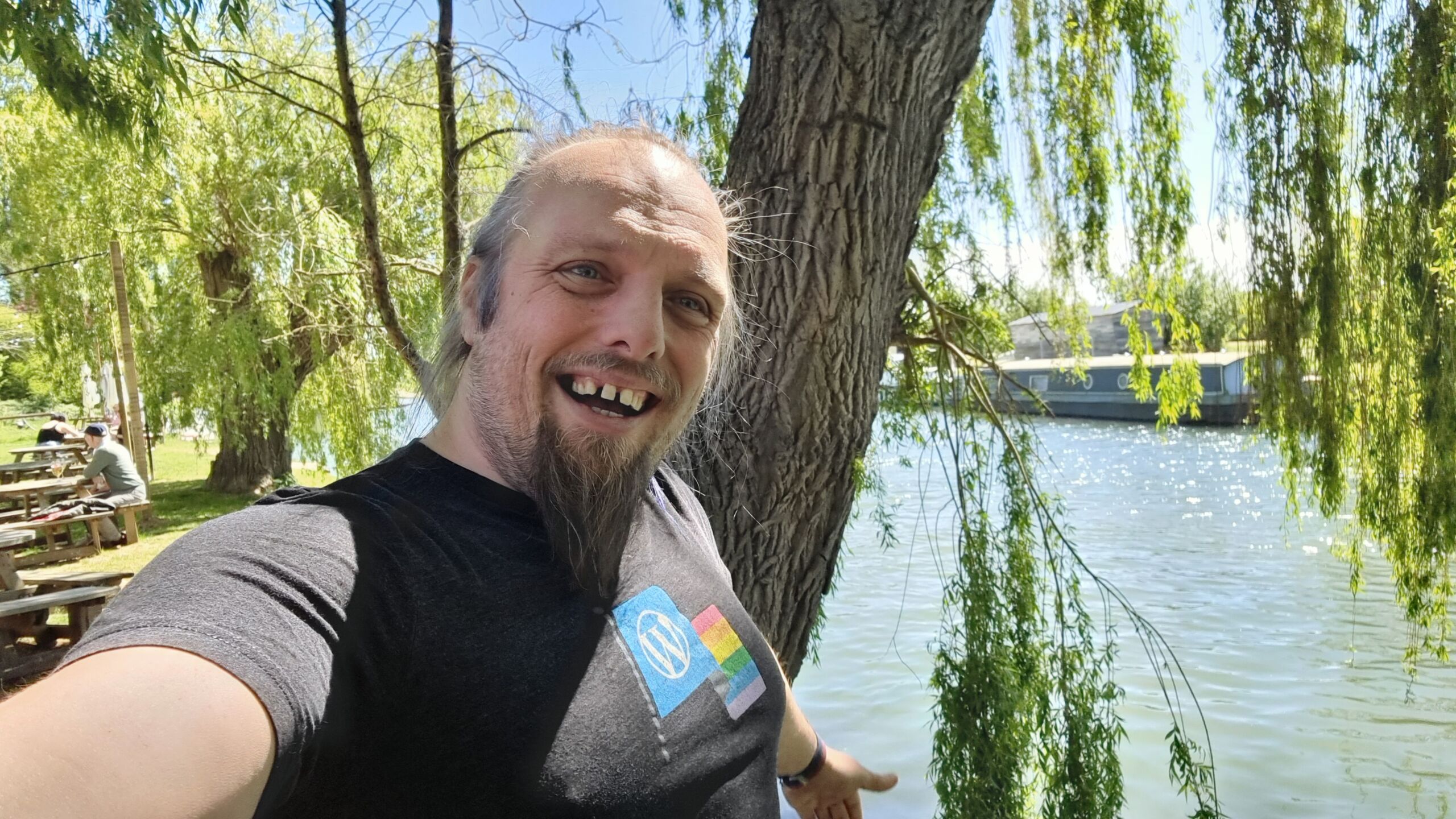 Full image of Dan, a white man with a blue ponytail and a goatee, smiles next to a broad rural river.