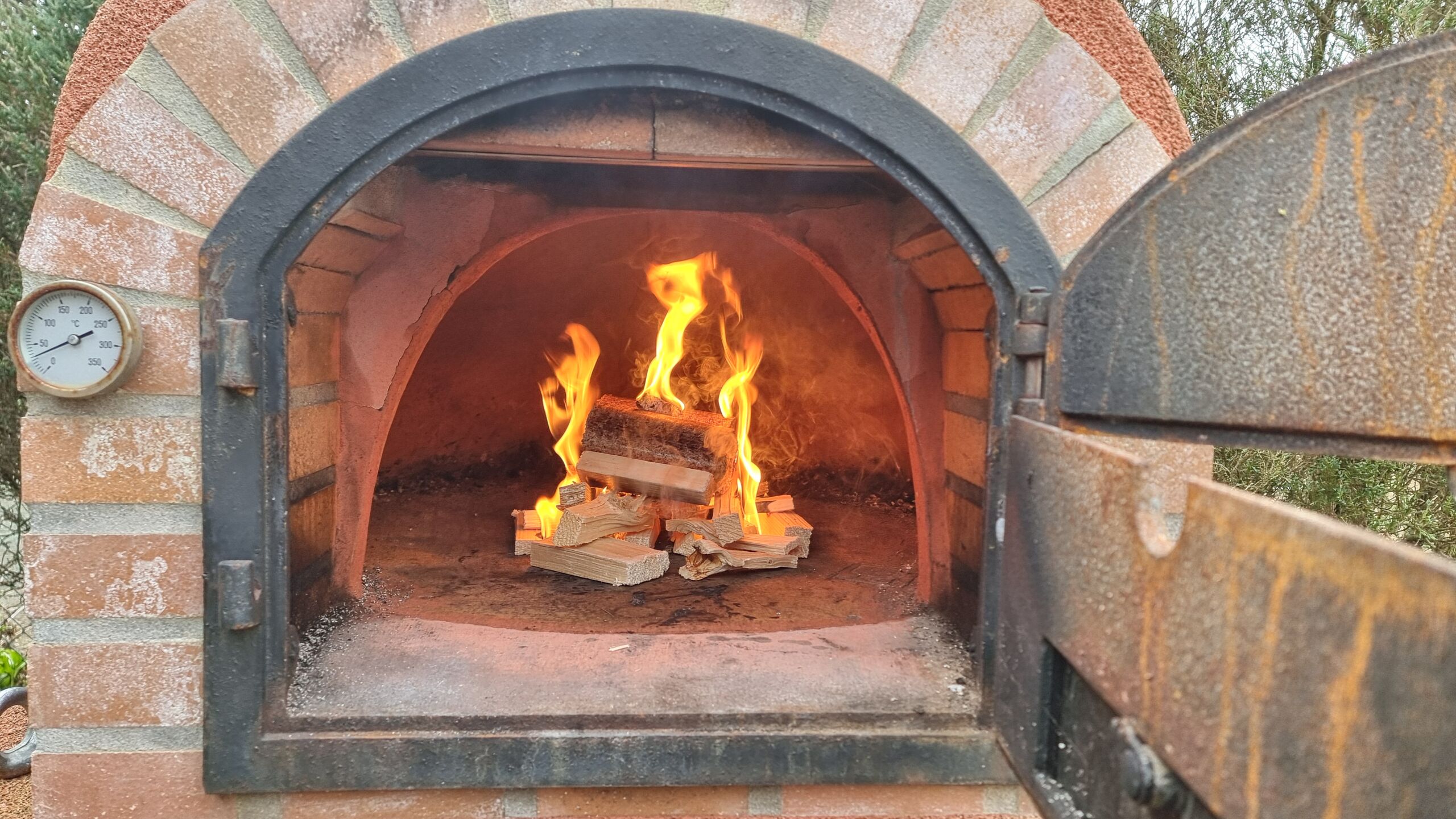 Full image of A small wood fire burns inside an outdoor brick pizza oven.
