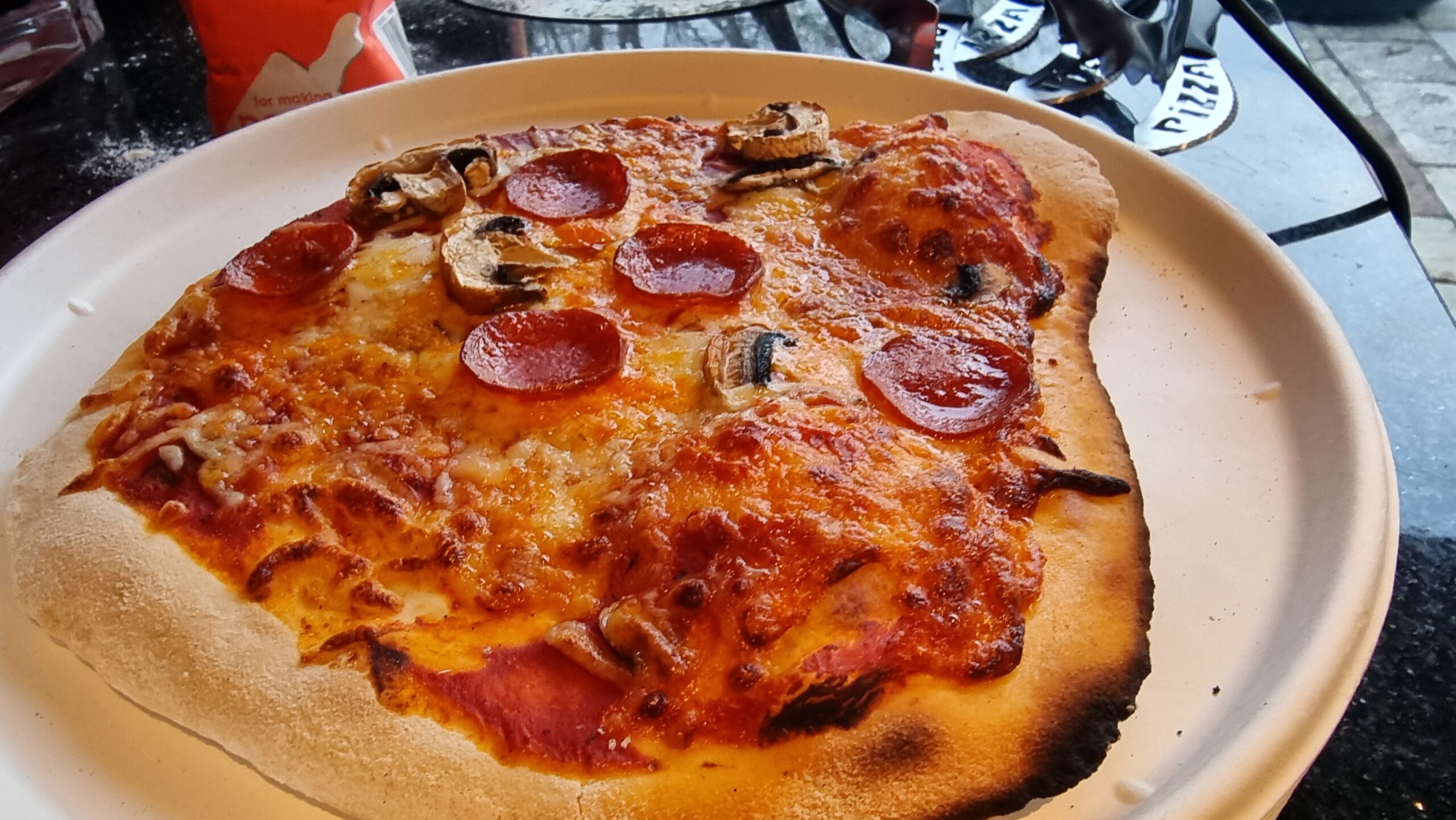 Full image of A crispy, misshapen, slightly charred pepperoni and mushroom pizza on a paper plate.