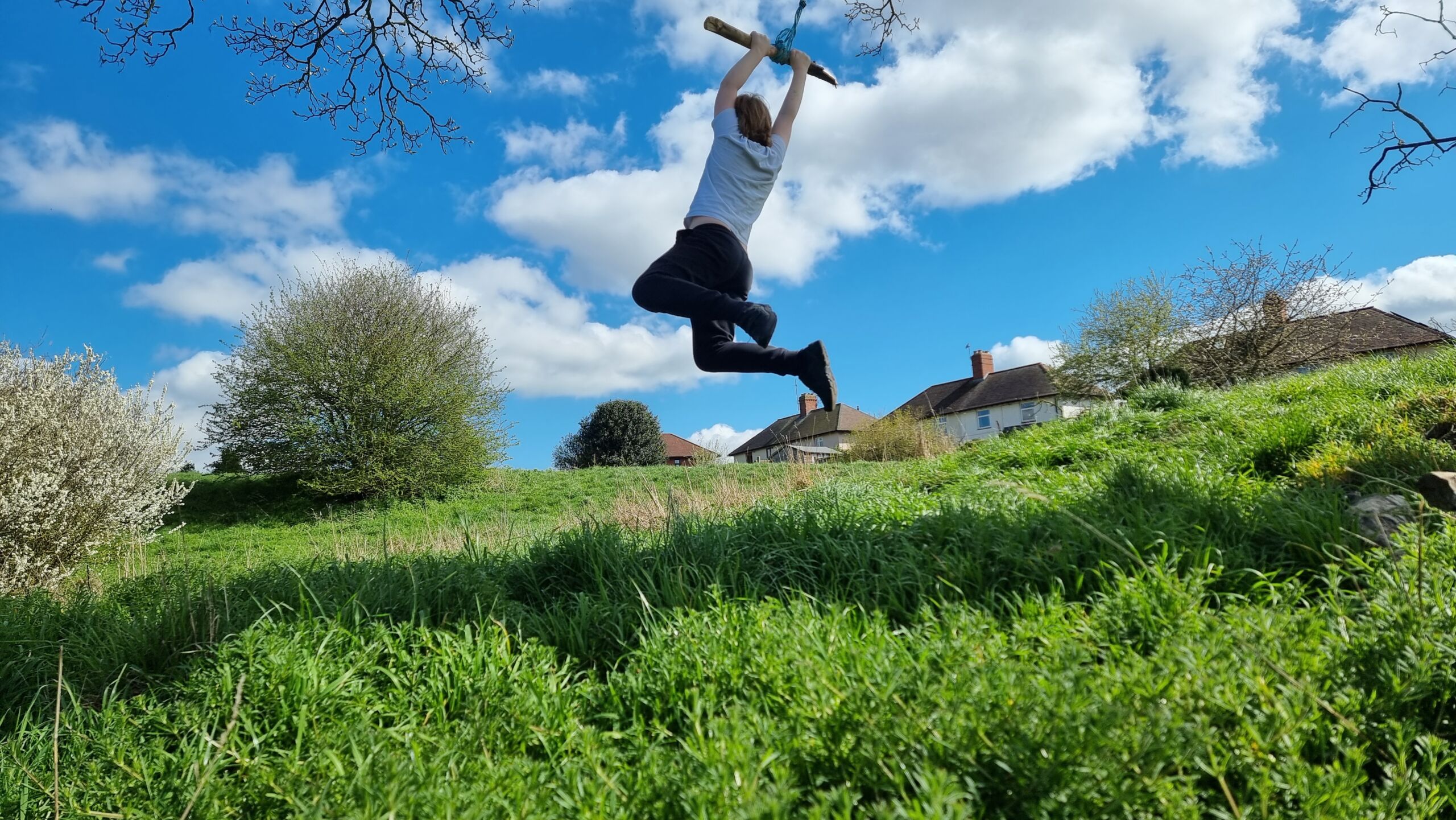 Full image of A boy hangs from a rope swing over a grassy hillside.
