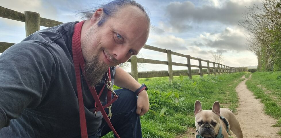 Dan crouches on a footpath running alongside a field, next to a French Bulldog.