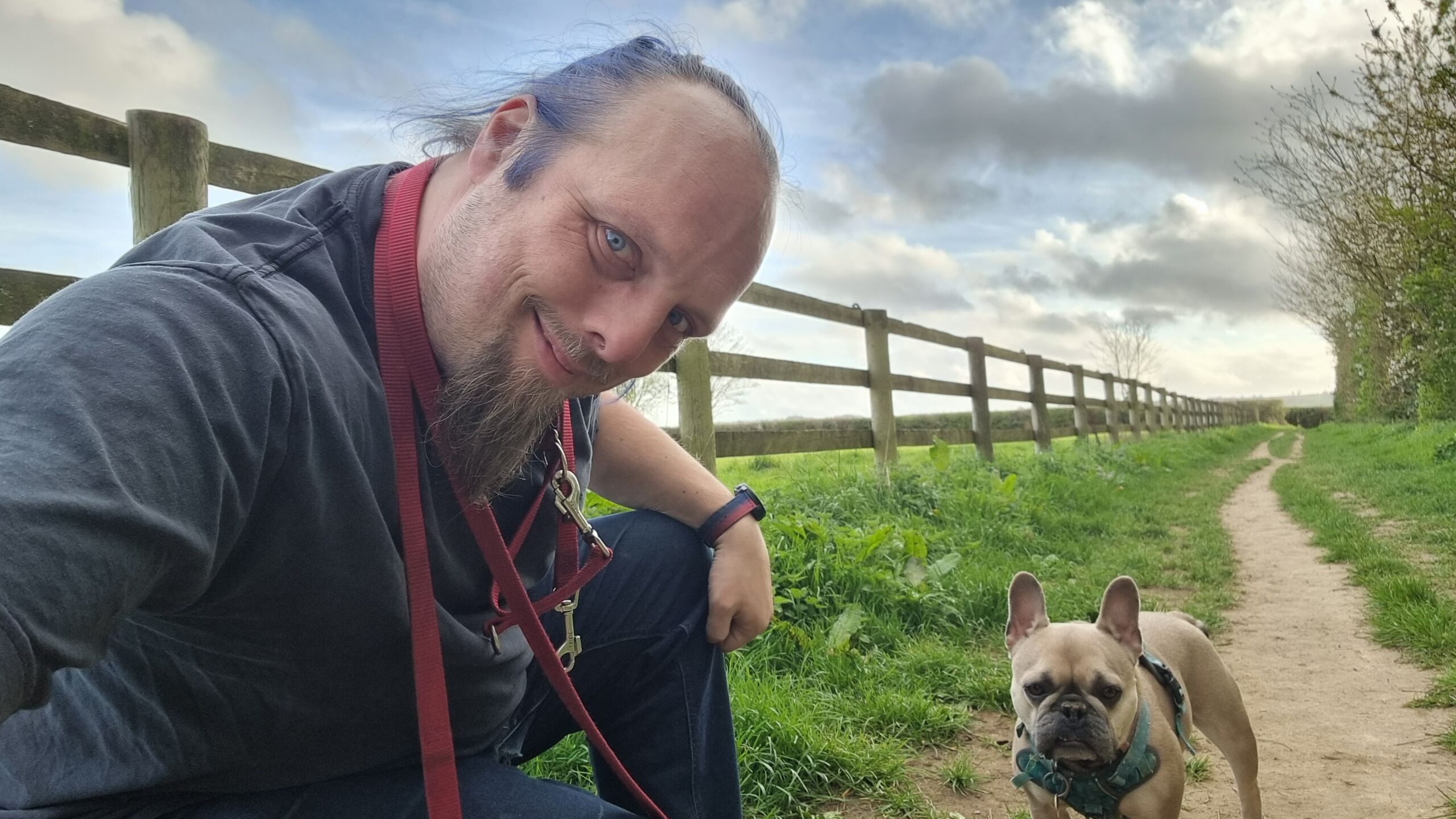 Full image of Dan crouches on a footpath running alongside a field, next to a French Bulldog.