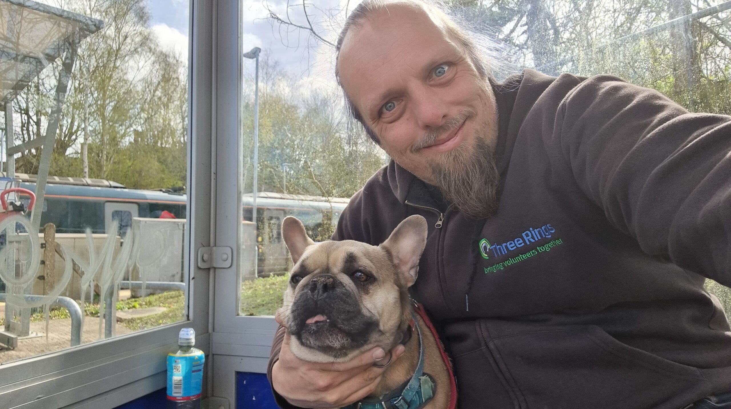 Full image of Dan sits in a glass bus shelter, hugging a French Bulldog.