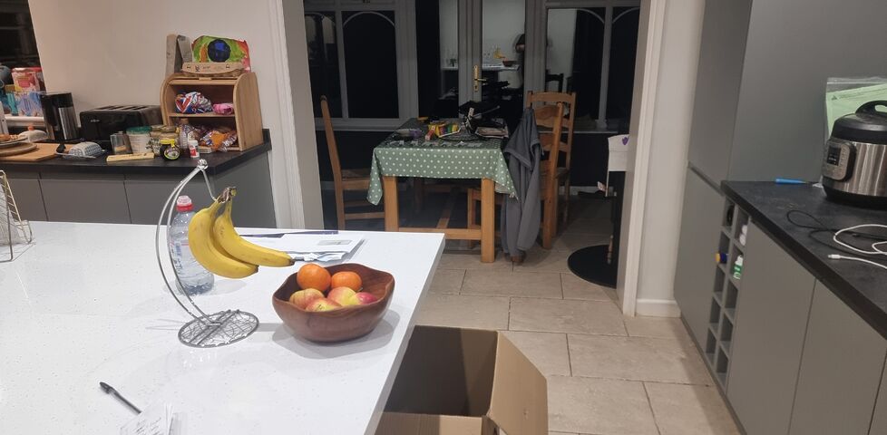 A moderately tidy kitchen with faux-marble countertops, under free on which an open large cardboard box can be seen. In the distance, a conservatory contains a small dining table cluttered with computer equipment.