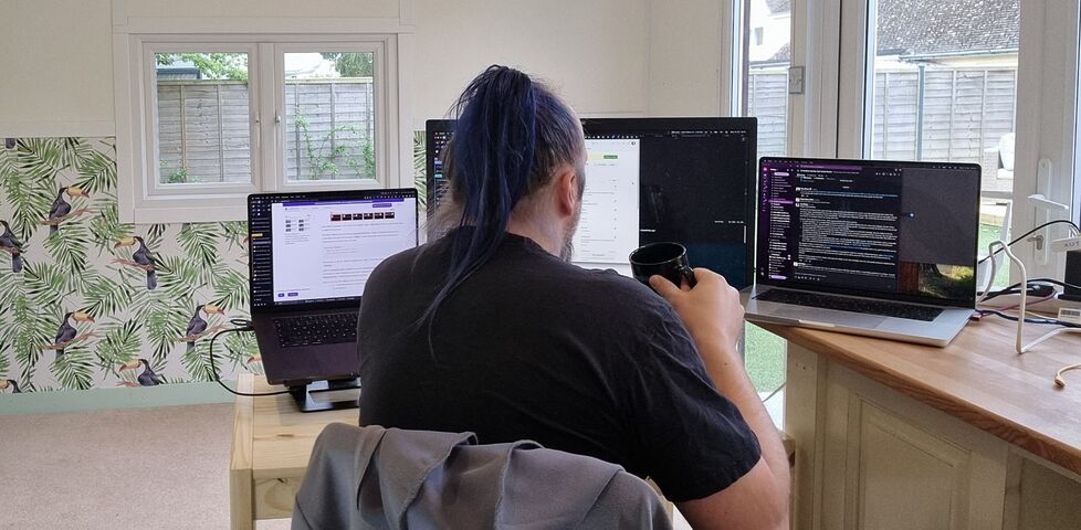 A white man with blue hair tied up in a ponytail sits at a basic pine desk in a garden office, decorated with wallpaper showing toucans. In front of him are two laptops and a large monitor. He holds a black mug in his hand, as if about to drink from it.
