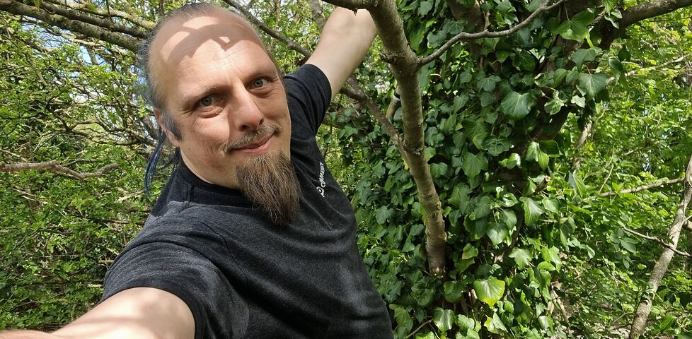 Dan hangs from a tree; a cache container is barely visible in the background.