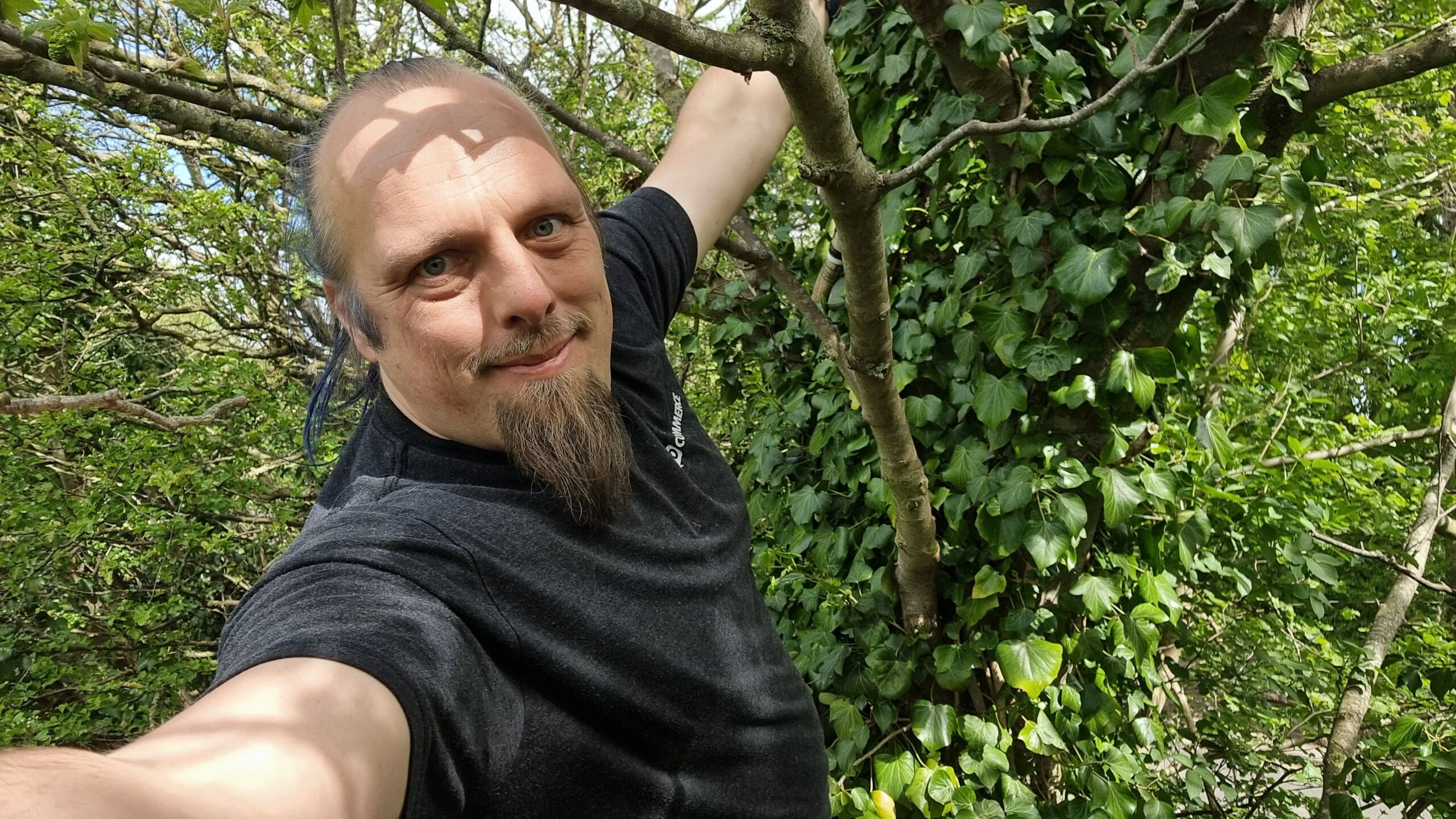 Full image of Dan hangs from a tree; a cache container is barely visible in the background.