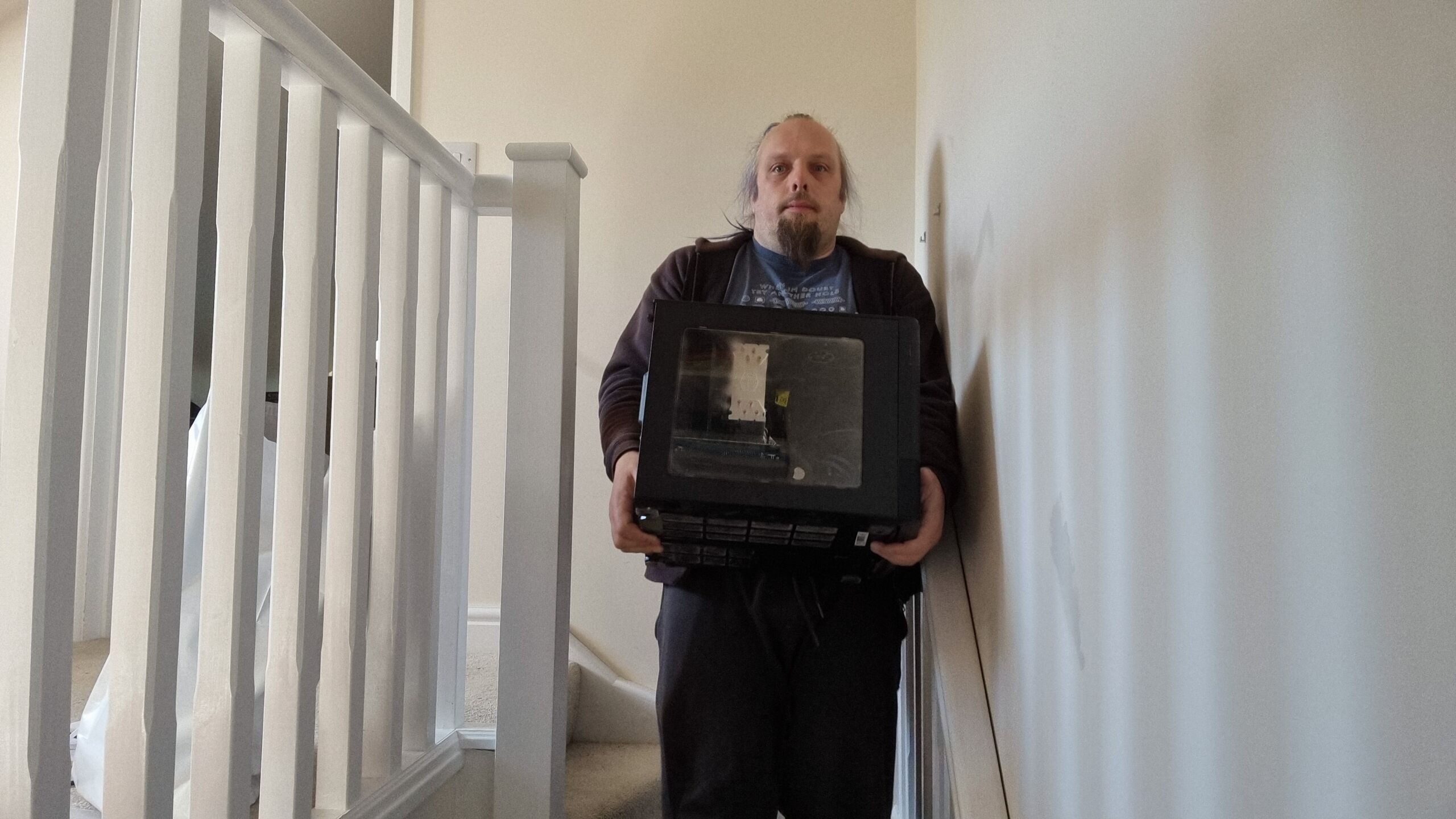 Full image of Dan, a white man with a ponytail and a goatee beard, carries a large black cube-shaped computer down a staircase.