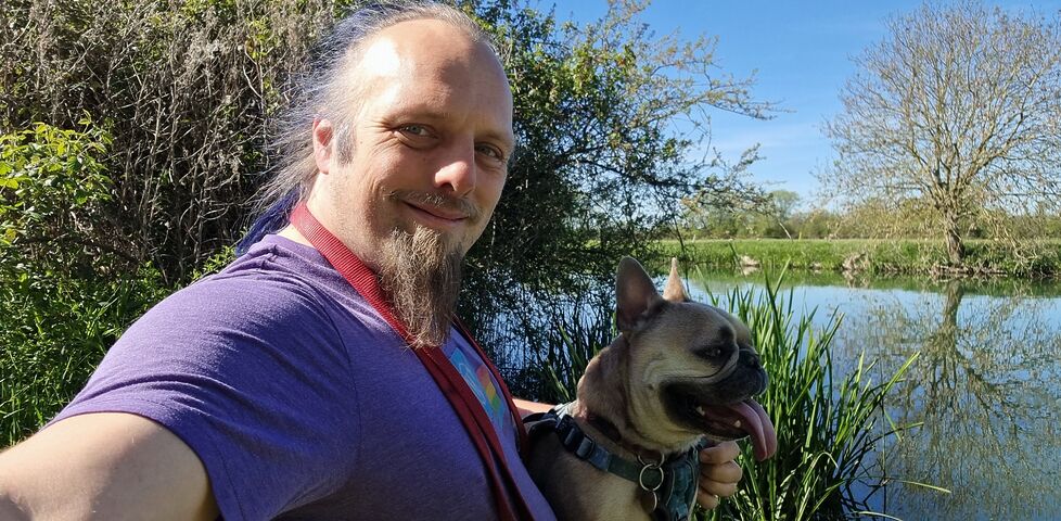 A white man with a blue ponytail and a goatee beard sits with a French bulldog alongside a wide, calm river.