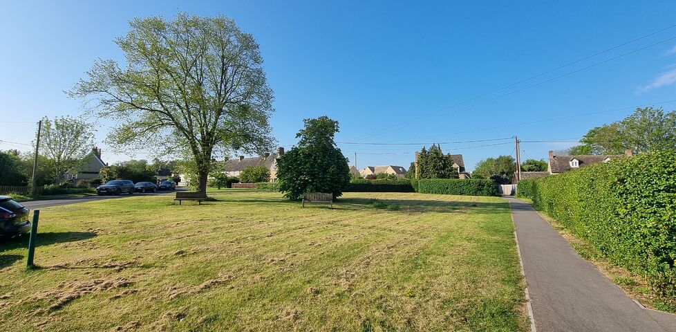 A village green with trees and benches under clear blue skies and Spring morning sunlight.